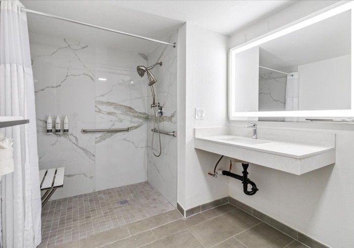 An accessible bathroom featuring a white marble-tiled walk-in shower with grab bars and a floating vanity with a lit mirror.