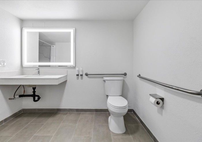 A modern, accessible bathroom with a white sink, an illuminated mirror, a toilet with grab bars, and light-colored tile.
