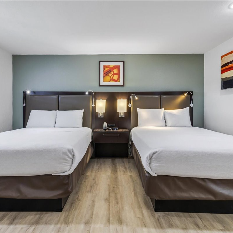 A hotel room featuring two queen beds with white linens, dark brown headboards, and a shared nightstand against a teal wall.