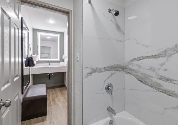 A view of a modern bathroom with a white marble-tiled bathtub and a doorway leading to a vanity area with a mirror.