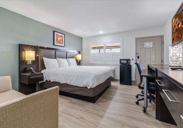 A hotel room with a queen bed, light wood floors, a desk with a chair, a mini-fridge, and a beige armchair.