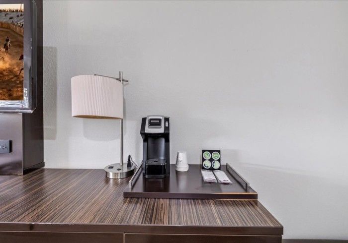 A coffee maker, a lamp with a white shade, and coffee pods on a tray, all sitting on a dark wooden desk.