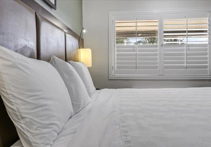 A made hotel bed with white linens and pillows against a dark brown headboard, next to a window with white shutters.