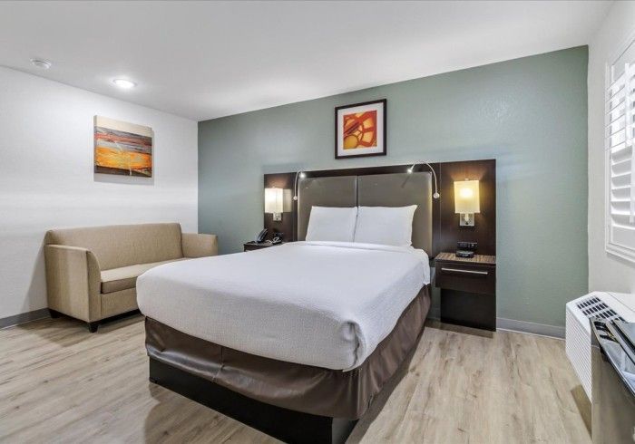 A hotel room with a queen bed, a tan sofa, light wood-look flooring, and a green accent wall with a framed print.