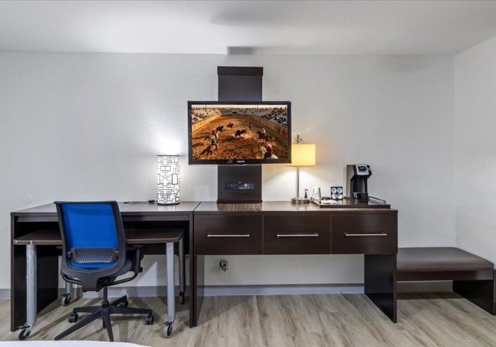 A hotel room desk area with a blue office chair, a flat-screen TV mounted on the wall, a lamp, and a coffee maker.