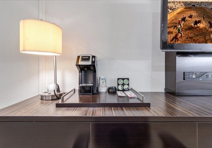 A hotel desk featuring a table lamp, a coffee maker on a tray, and a television screen showing a horse arena.