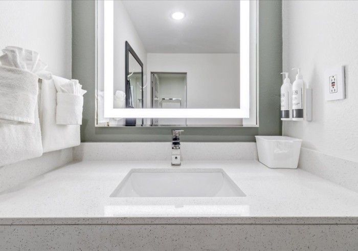 A clean hotel bathroom vanity with a white countertop, rectangular sink, illuminated mirror, and neatly folded towels.
