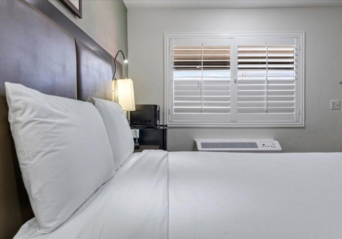 A hotel bedroom with a large bed, two white pillows, a headboard, a bedside lamp, and a window with white shutters.