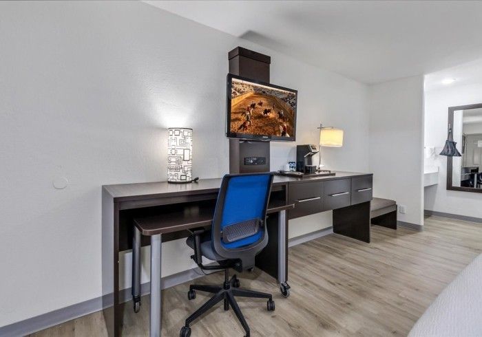 A hotel room workspace with a desk, a blue office chair, a television mounted on the wall, and a lamp on wood flooring.