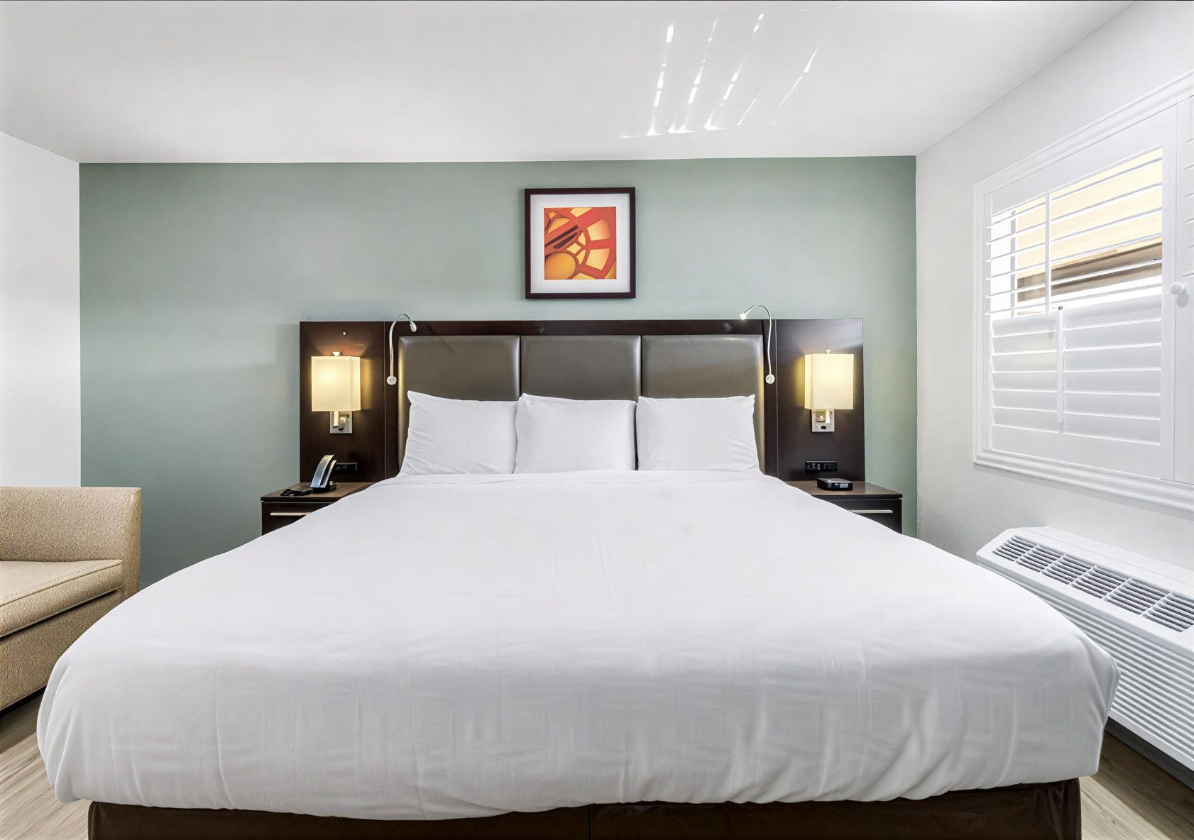 A hotel room with a queen bed featuring a white comforter, two bedside lamps, a framed wall print, and a side chair.