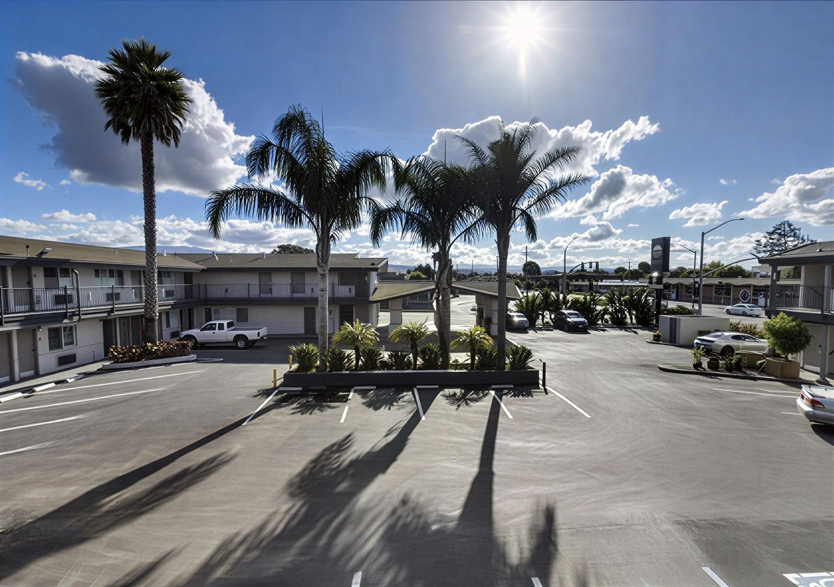 A parking lot in front of a motel building with palm trees under a sunny blue sky with clouds.