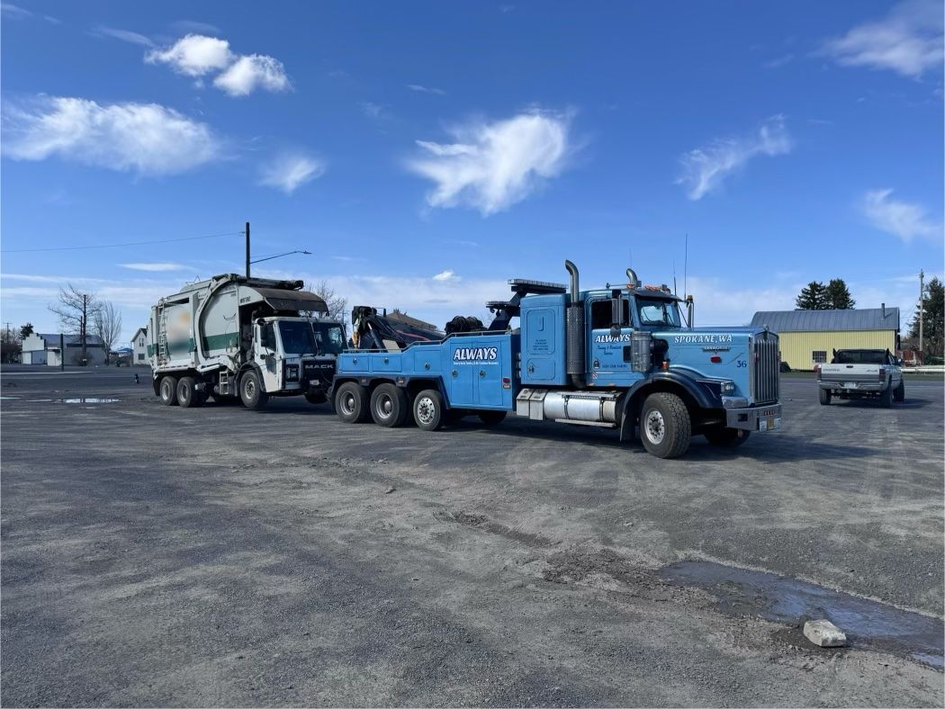 A blue tow truck is towing a garbage truck in a parking lot.