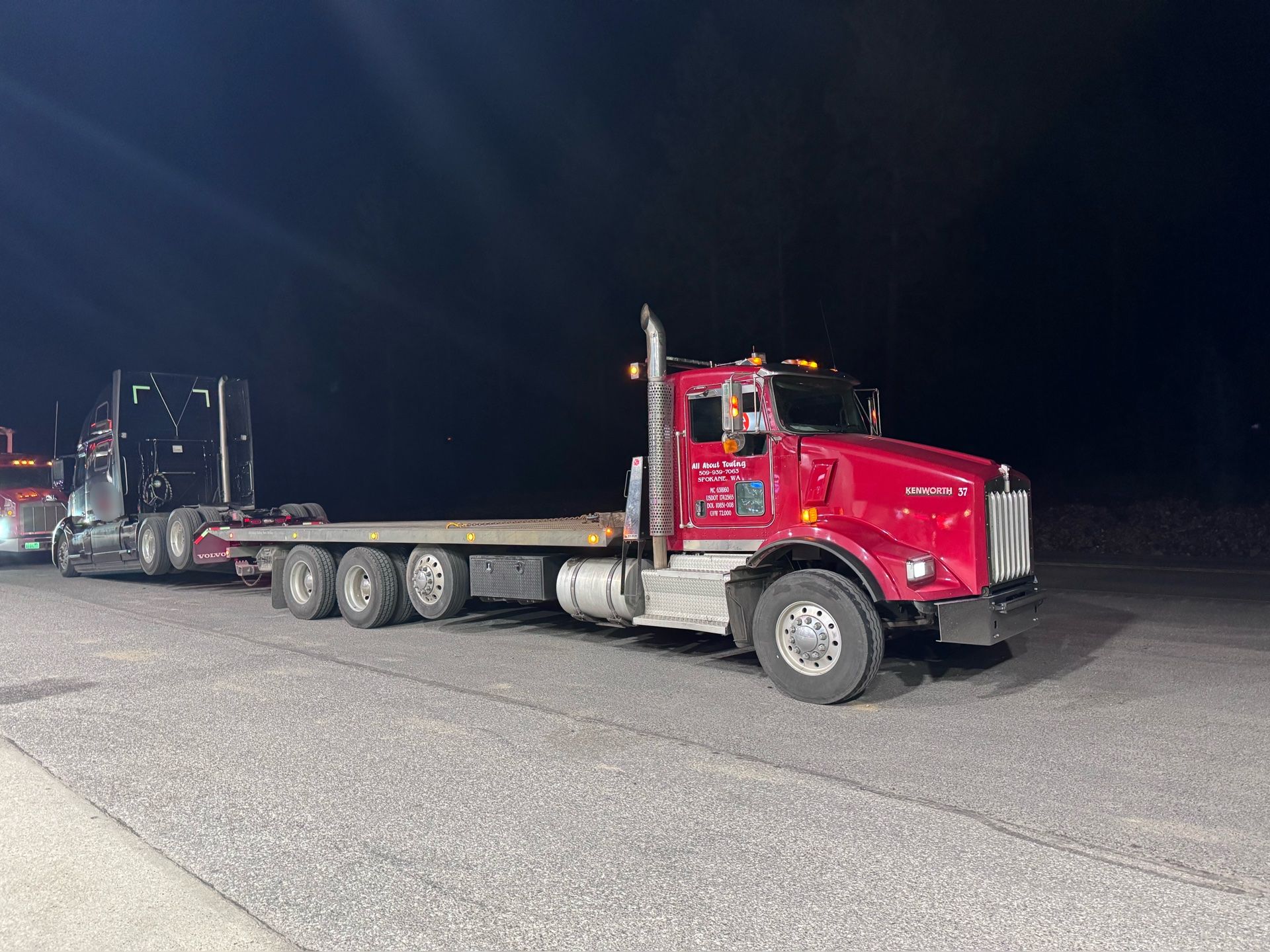 A red semi truck with a flatbed trailer is parked on the side of the road at night.
