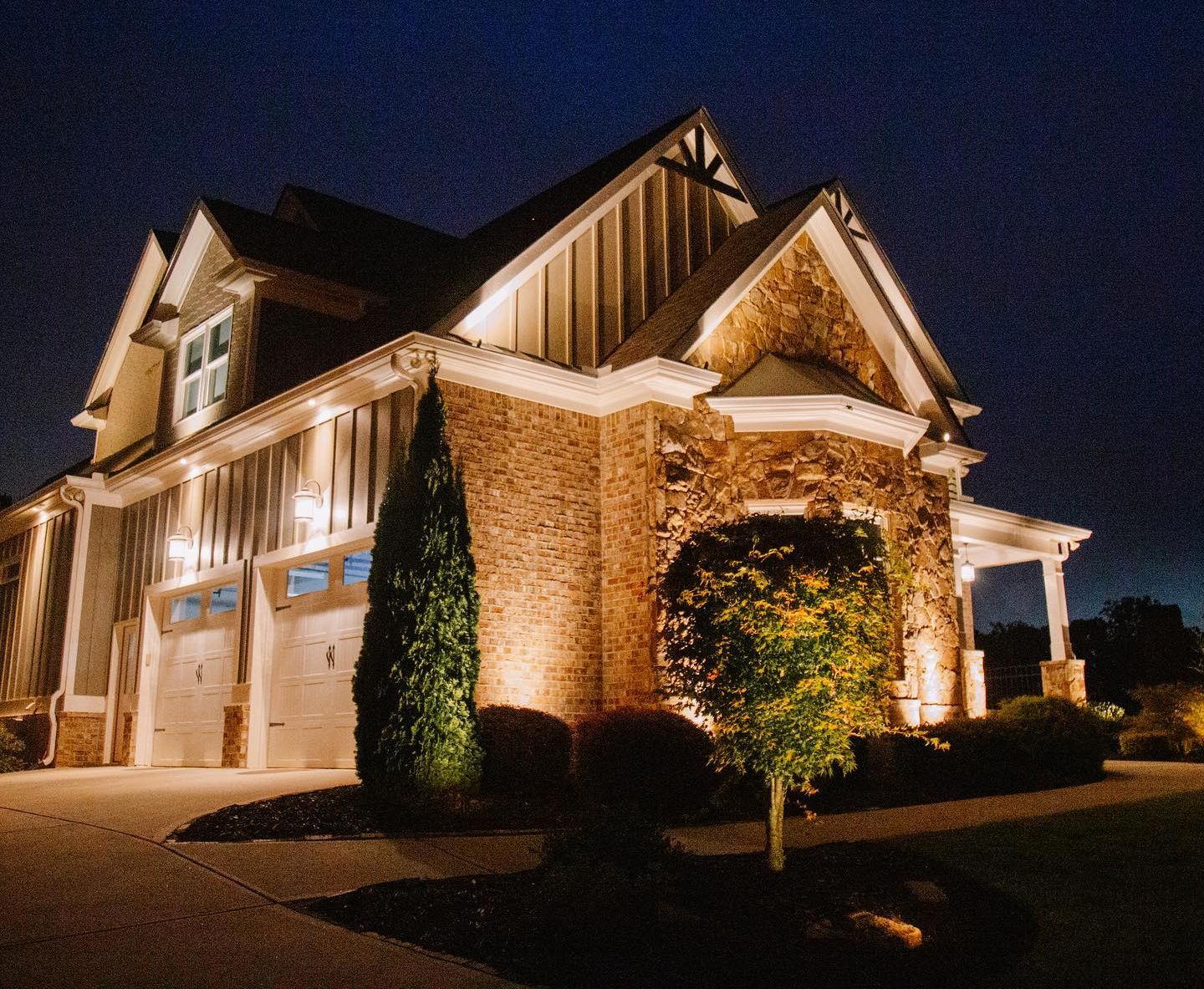 Two-story house illuminated at night. Brick and siding exterior, garage doors, front yard with trees, dark sky.