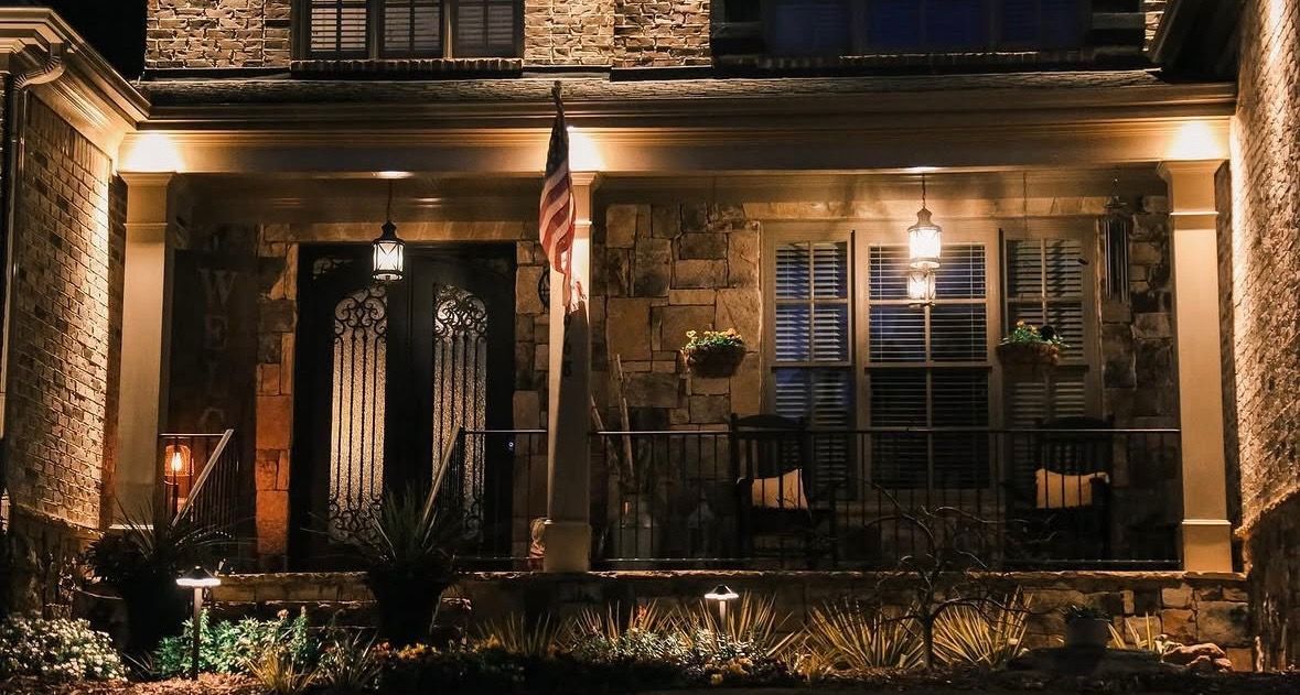 Warmly lit brick townhouse facade at night with porch columns, shuttered windows, and front garden lights