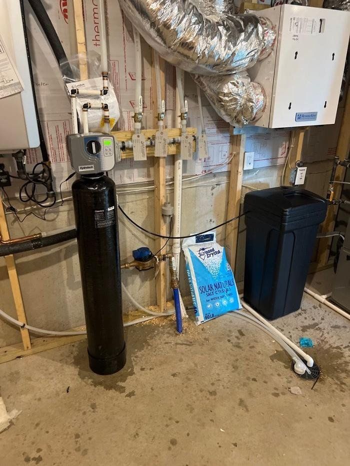 A water softener is sitting in a basement next to a trash can.