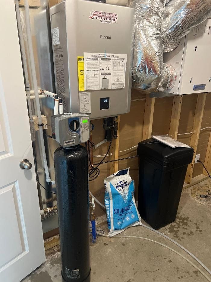 A boiler is sitting on top of a black tank in a room next to a trash can.