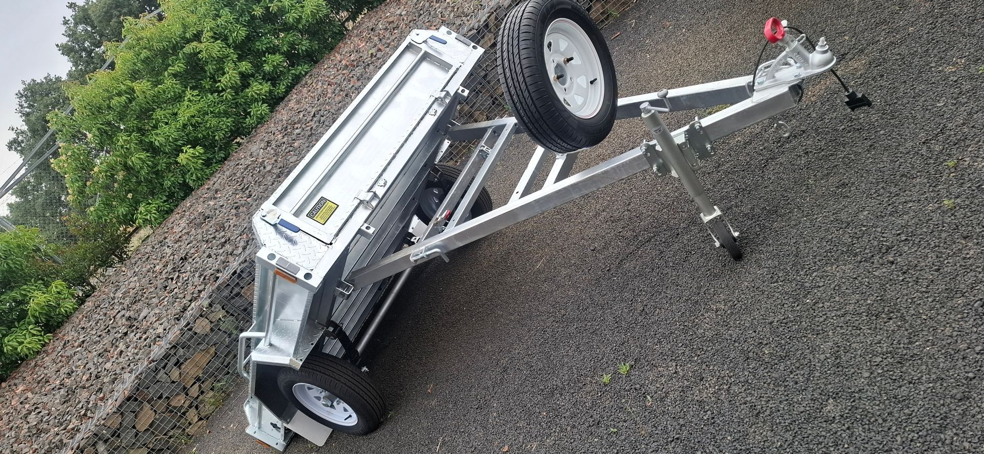 Silver utility trailer on gravel with tires and green foliage in background — Towform Trailers in Brocklehurst, NSW