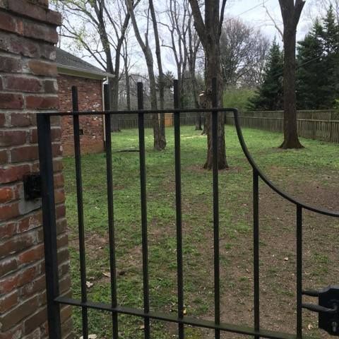 Black iron gate opens to a grassy yard with trees and a brick building.