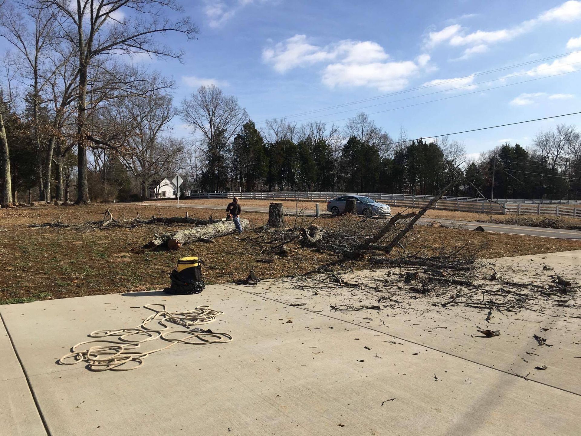 Man clearing debris on a lot with bare trees, small car, and house in the background, under a blue sky.