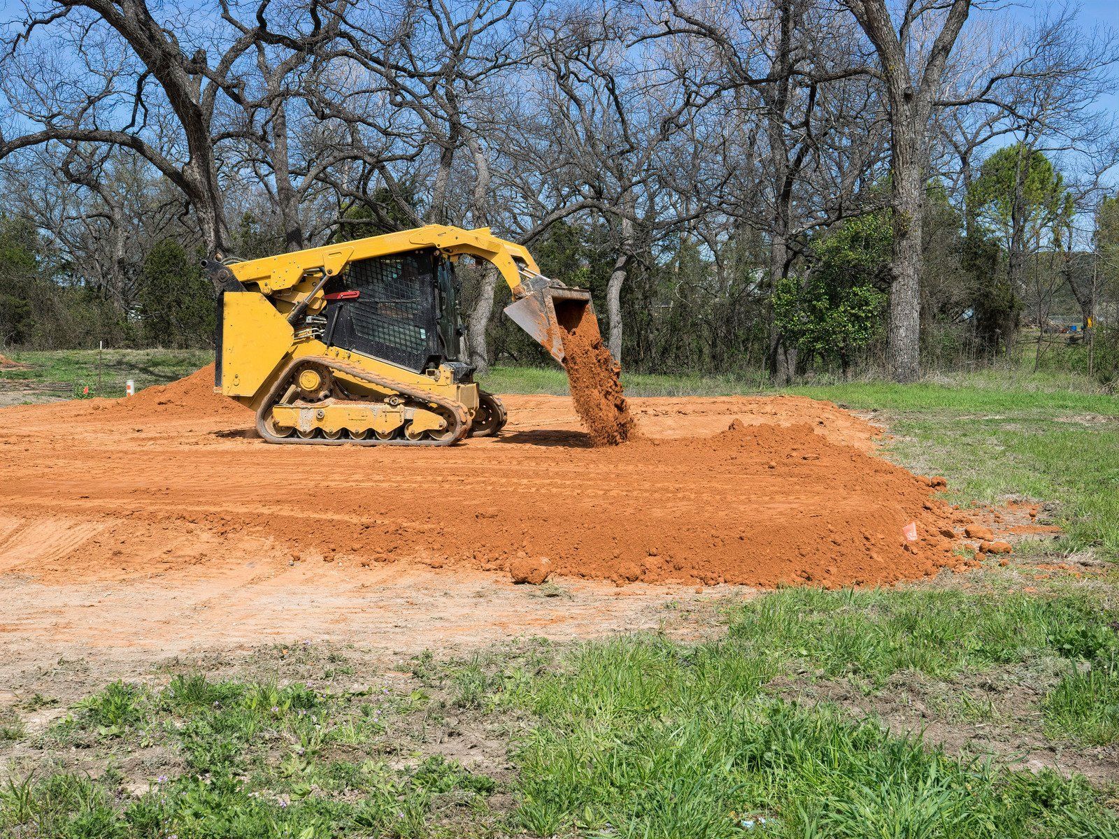 Yellow skid steer dumping dirt on a mound in a grassy field near trees.