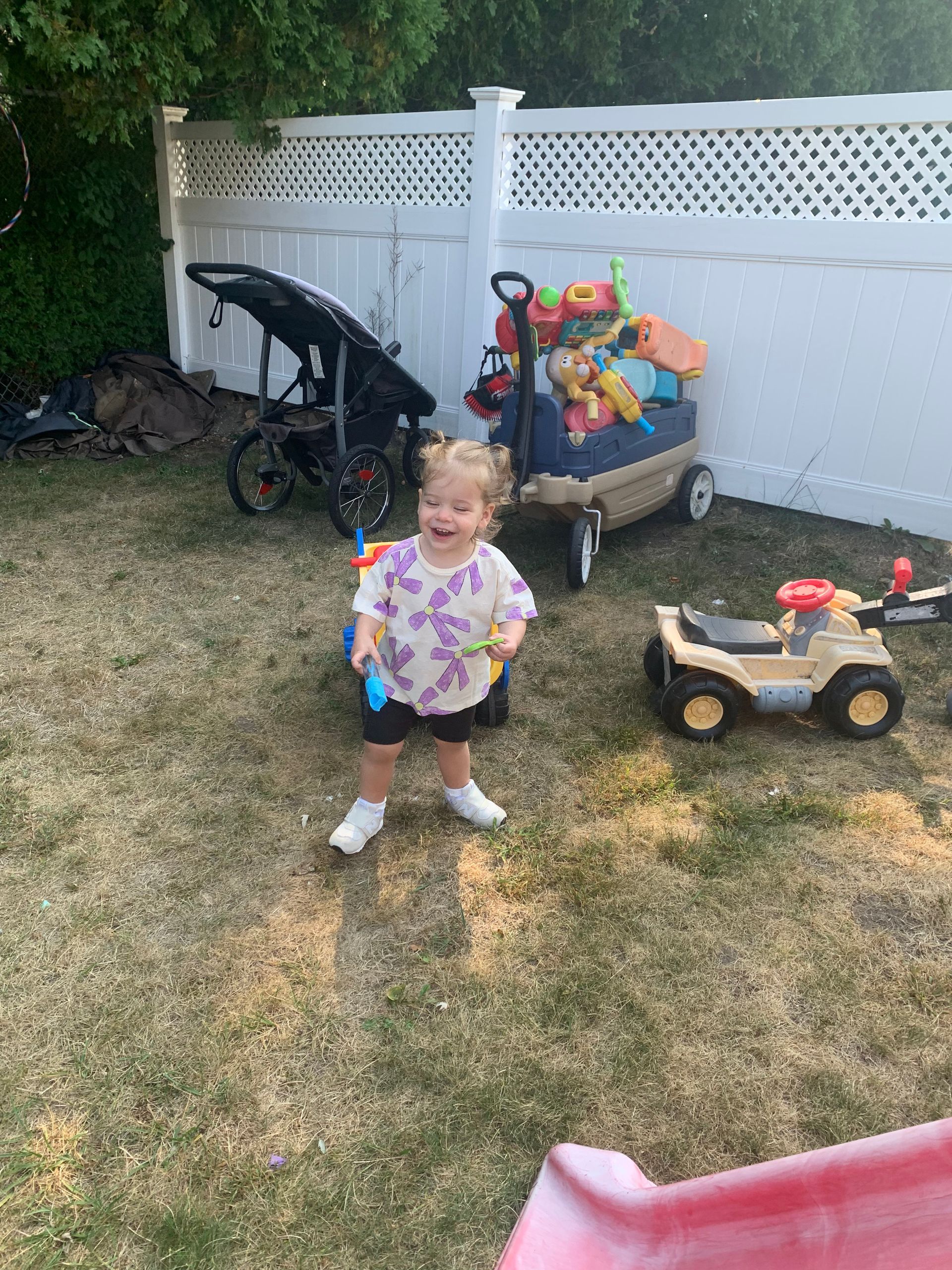 A little girl is standing in a backyard next to a wagon filled with toys.