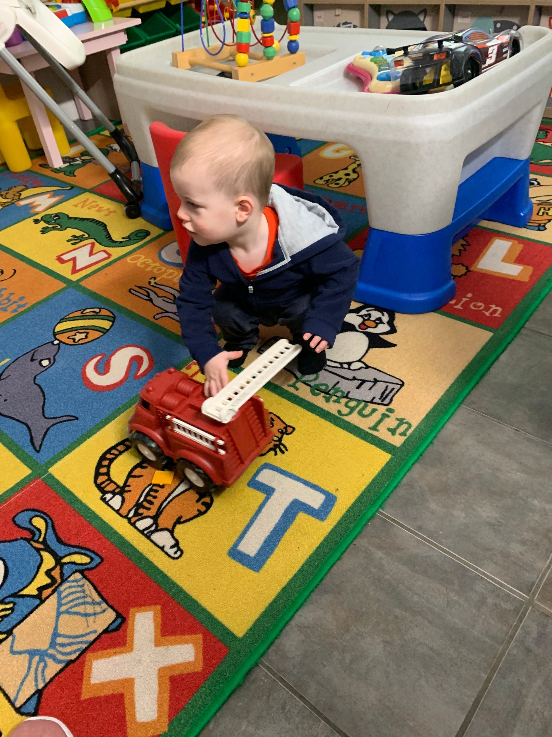 A little boy is playing with a toy truck on a rug with the letter t on it.