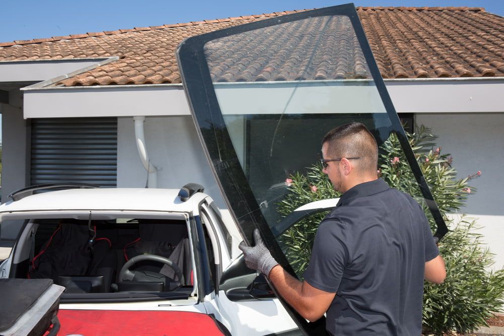 A Man is Installing a Windshield on a Car in Front of a House — Bowen Mechanical & Haulage in Bowen, QLD