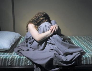 A woman is sitting on a bed with her head on her knees.
