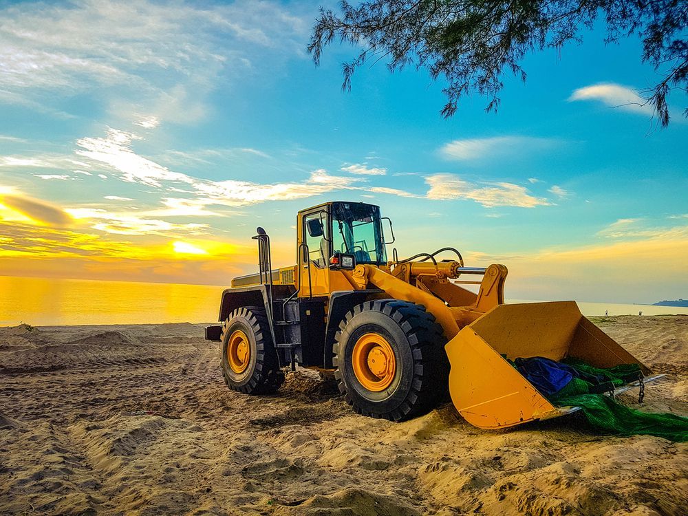 A Yellow Bulldozer Is Parked on The Beach at Sunset — PBC Compliancing Pty Ltd in Moranbah, QLD