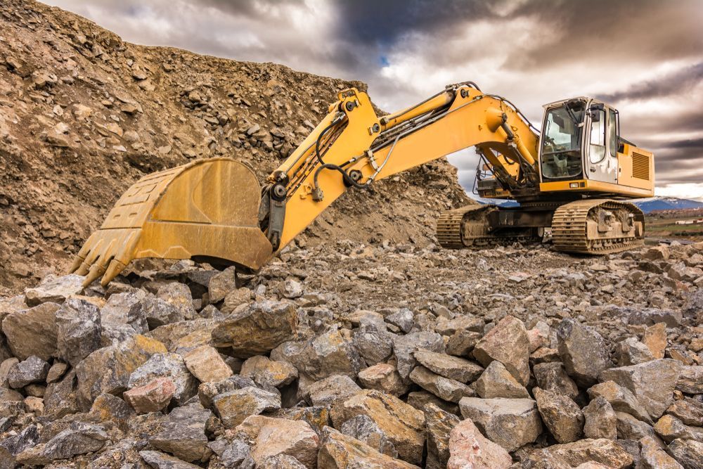 Yellow Excavator Working on a Pile of Rocks at a Quarry Under a Cloudy Sky — PBC Compliancing in Dysart, QLD