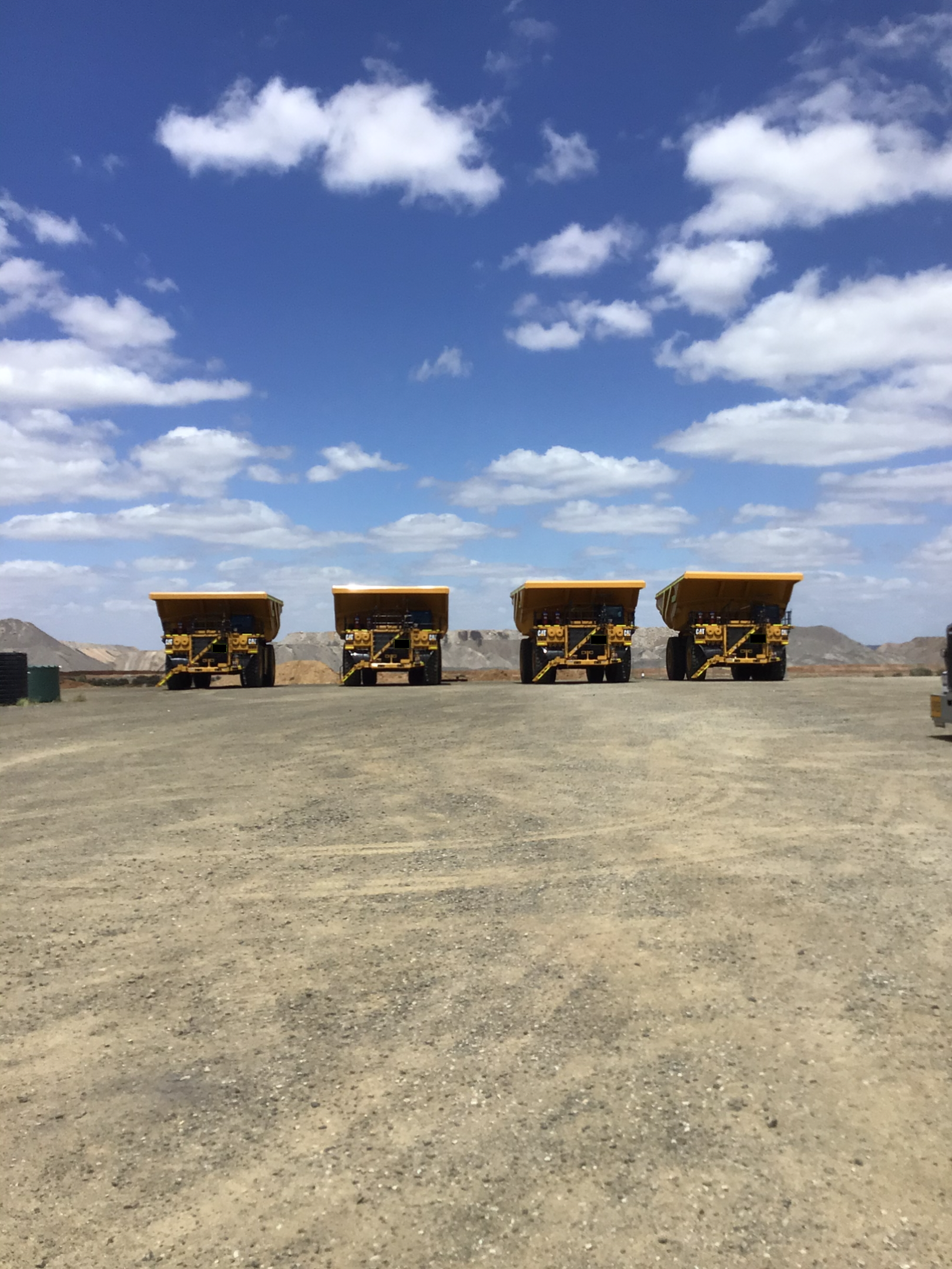 Four Yellow Mining Trucks Parked on A Dirt Lot Under a Blue Sky with Scattered Clouds — PBC Compliancing in Whitsundays, QLD