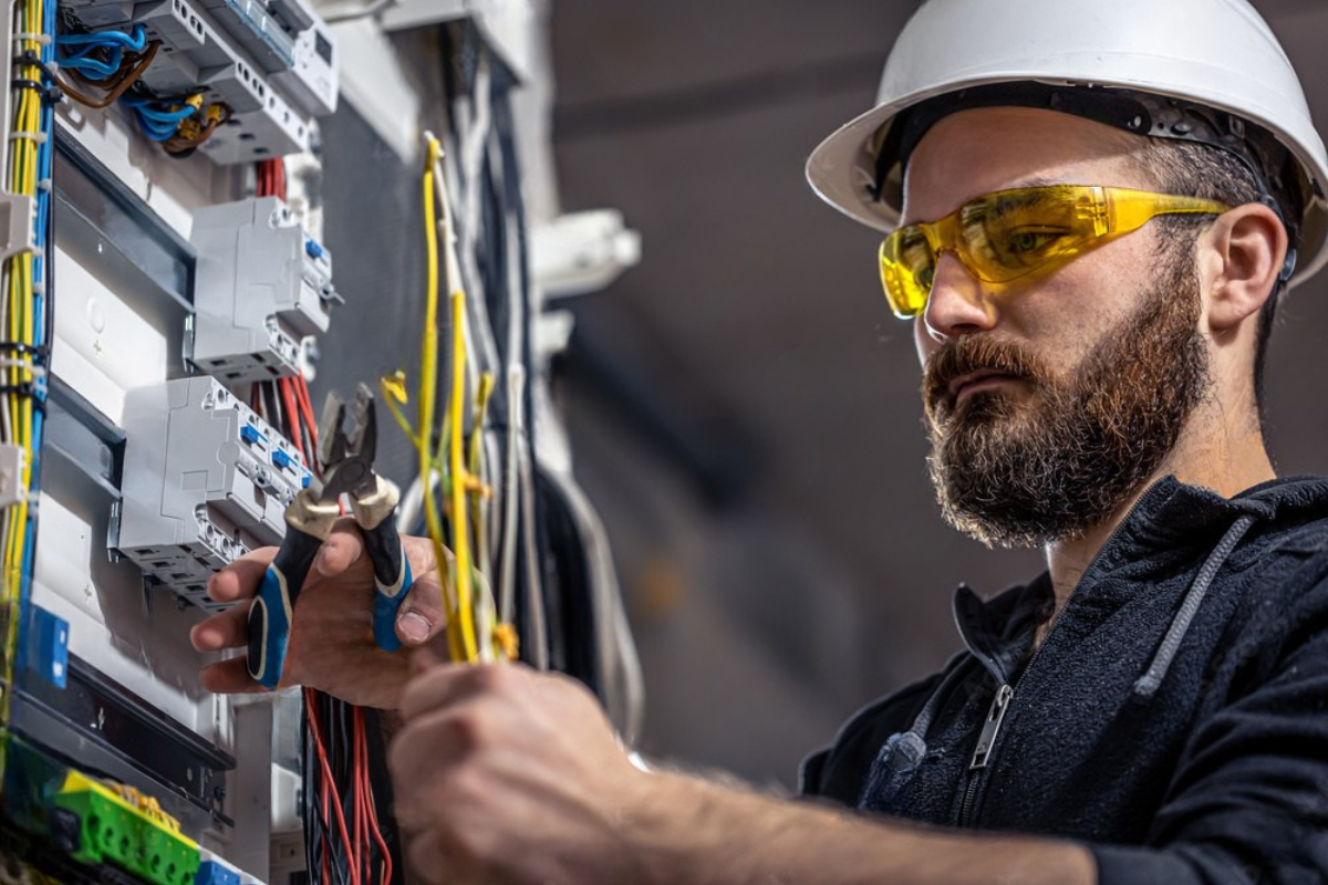 Electrician in hard hat and safety glasses working on wiring in an electrical panel.