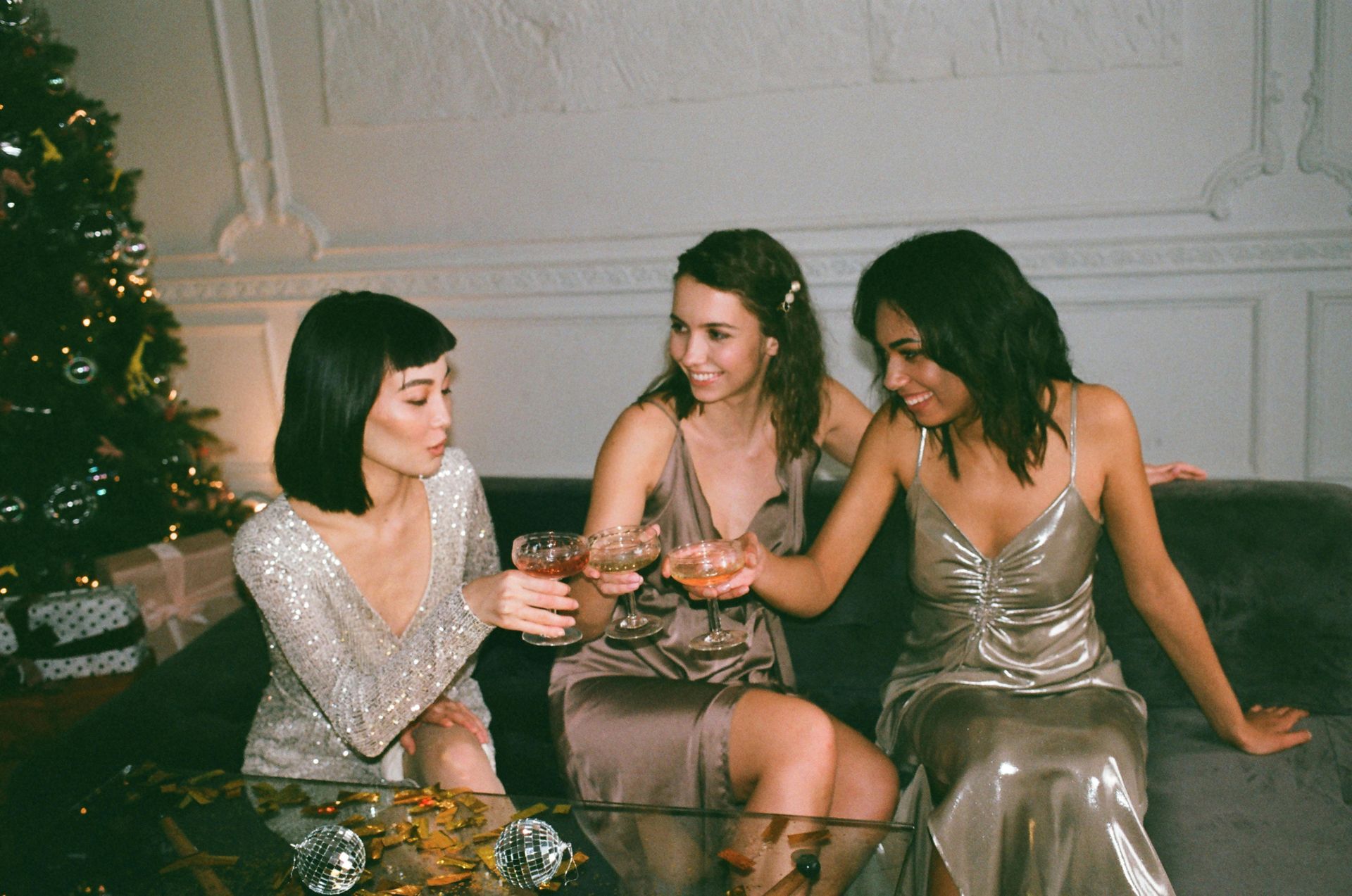 Three women in sparkly dresses clinking glasses, celebrating on a sofa near a Christmas tree.