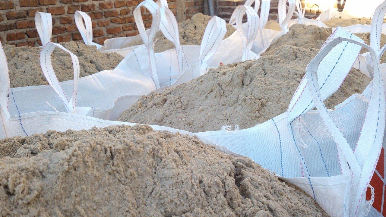 Grandes bolsas blancas llenas de arena, apiladas al aire libre contra una pared de ladrillos.