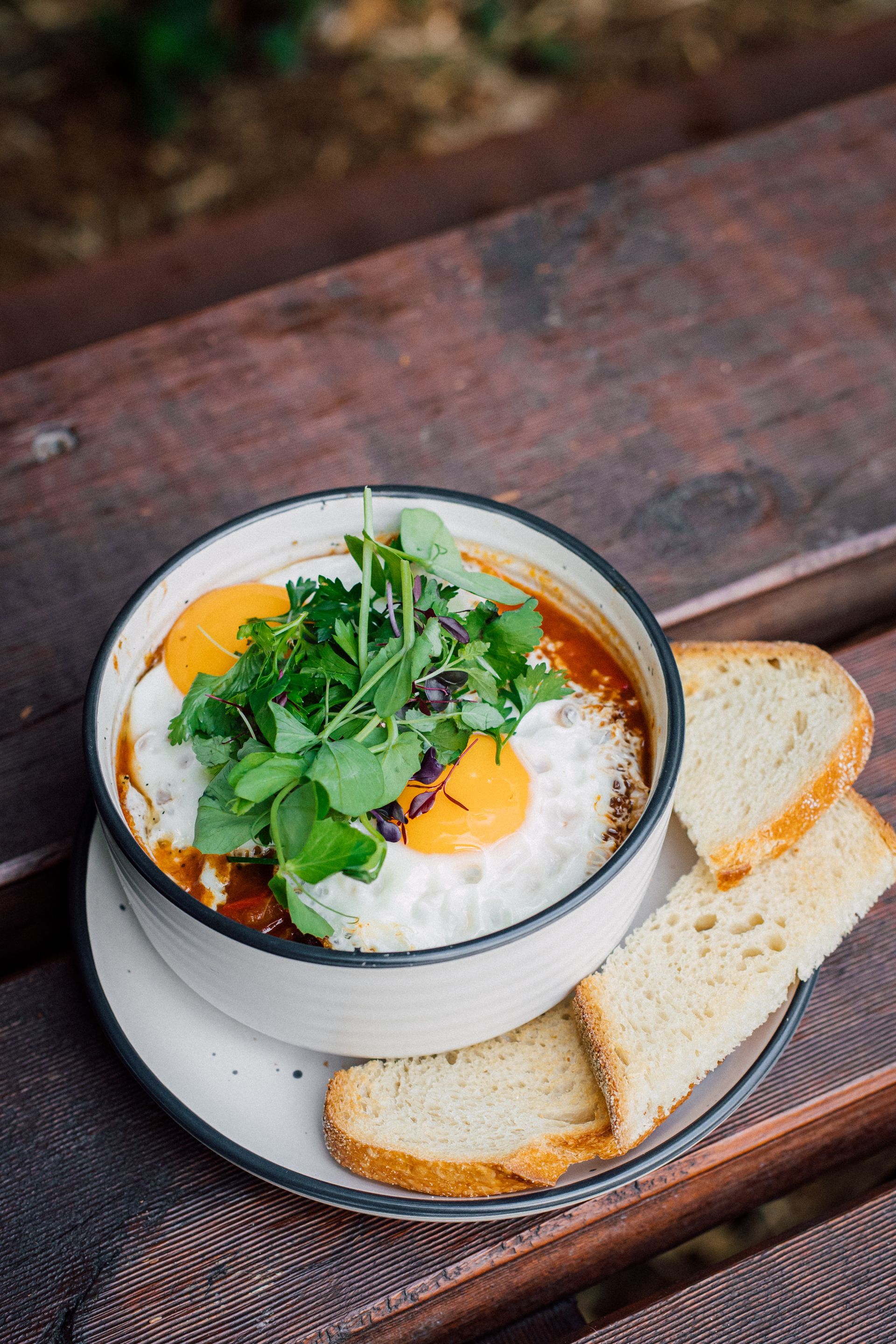 A bowl of soup with eggs and toast on a plate on a wooden table.