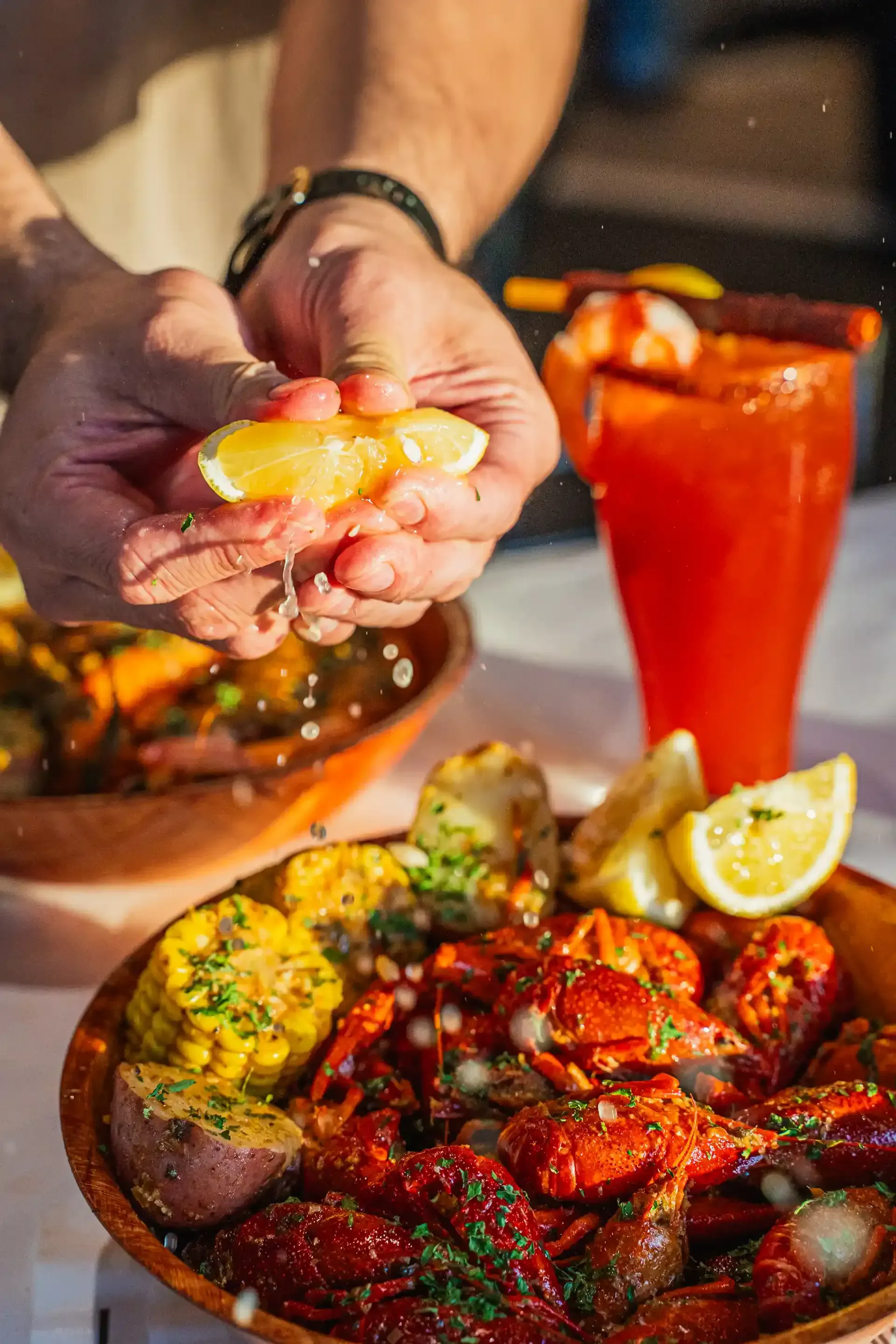 Hands squeezing lemon over a spicy seafood platter with a red drink and dipping sauce
