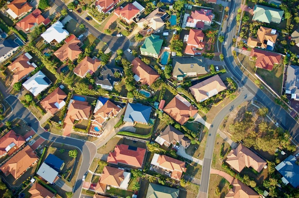 Aerial view of a suburban neighbourhood with houses, roads, driveways, pools, and trees.