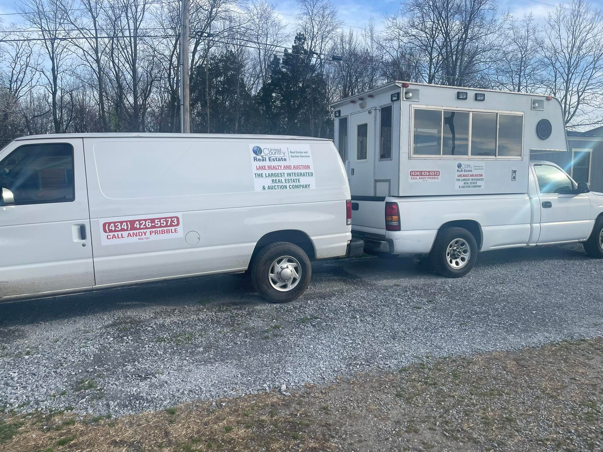 White van and pickup truck towing a camper. Both vehicles are parked on gravel, with trees in the background.