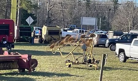 Agricultural equipment, including a yellow hay rake, on display outdoors near vehicles.