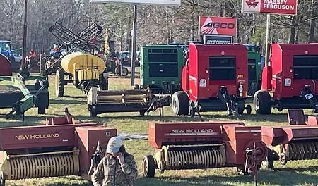 Farm equipment displayed outdoors, including tractors, balers, and sprayers; person visible in the foreground.