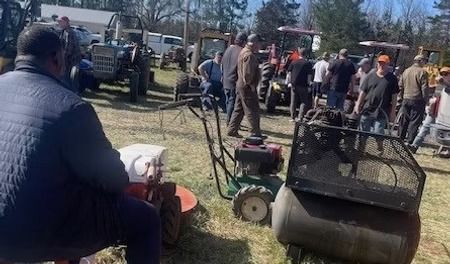 People at an outdoor auction with tractors, lawn mowers, and other equipment on display.