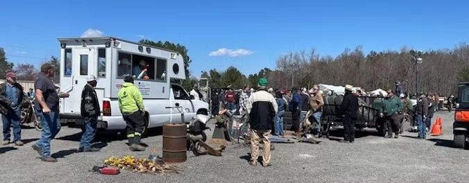 People gather in a parking lot near a white truck with a service box and a small vehicle, under a blue sky.