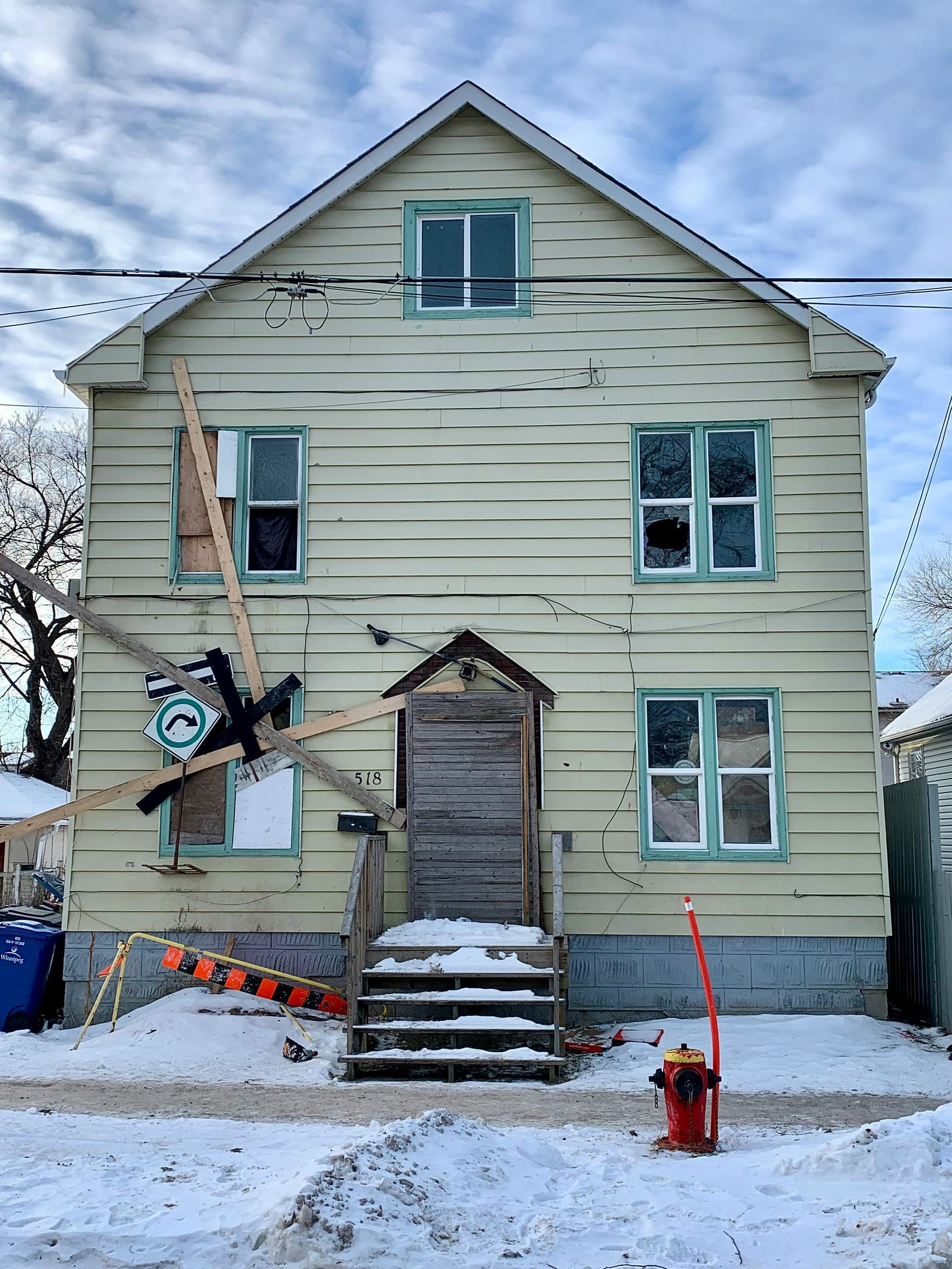 Damaged two-story house with boarded-up windows and an unusual object attached to the facade. Snow-covered ground.