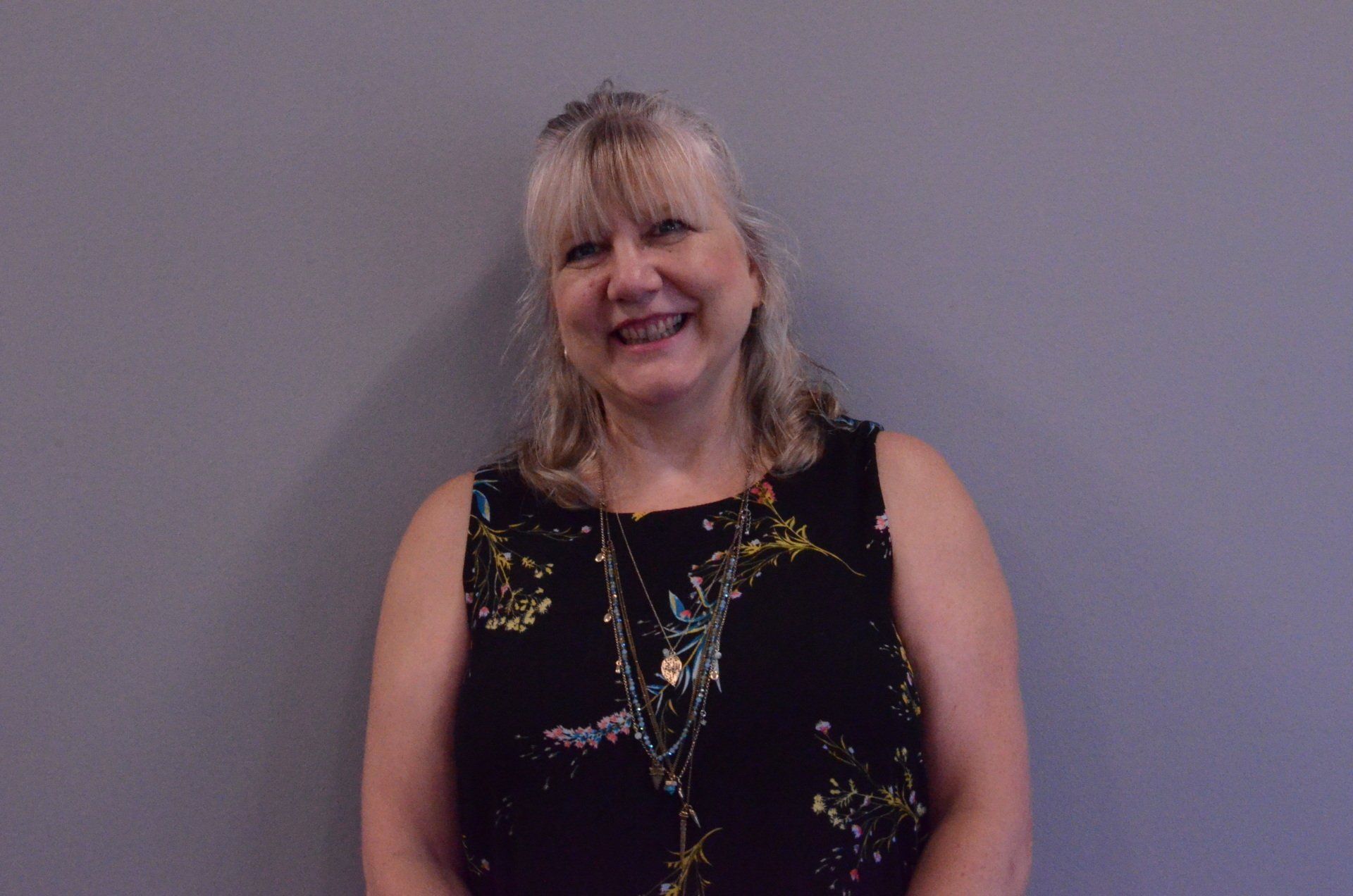 Woman in sleeveless floral top smiles in front of a gray wall.