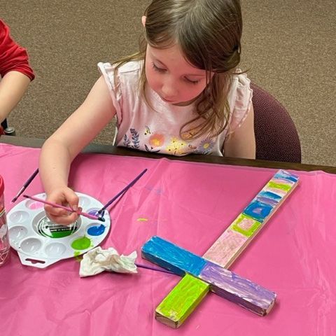 Girl painting a wooden sword with colorful paints at a table covered in pink paper.