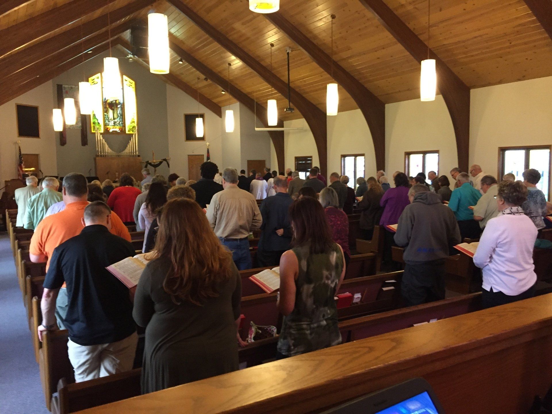 People standing in pews during a church service; arched wooden ceiling, stained glass window.