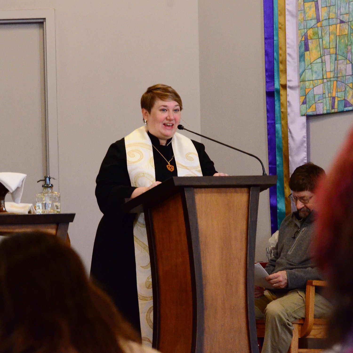 Woman in vestments speaks from a wooden podium. Smiling, with a microphone. In a light-filled room.