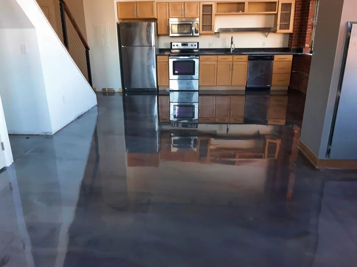 Shiny gray epoxy floor reflecting a kitchen with stainless steel appliances and light wood cabinets.