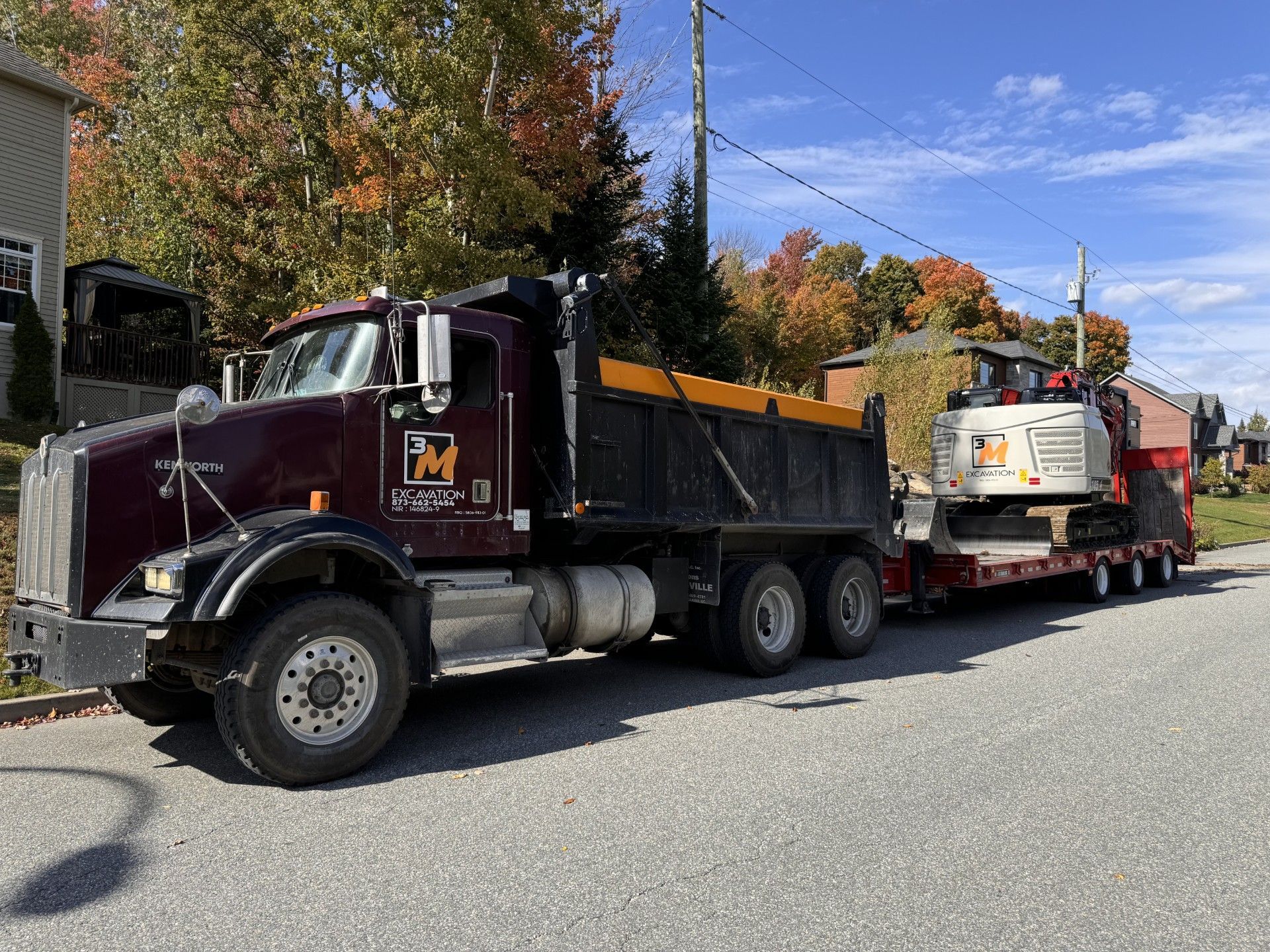 Camion à benne basculante rouge foncé avec une petite excavatrice sur une remorque garée dans une rue bordée d'arbres colorés.